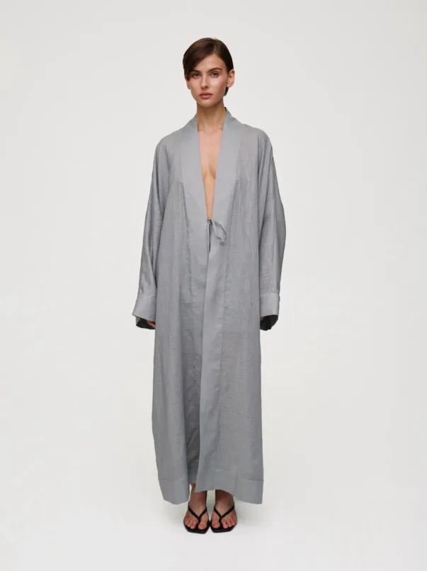 Model wearing a loose gray robe with a waist tie, standing against a pale background, wearing black strappy sandals.