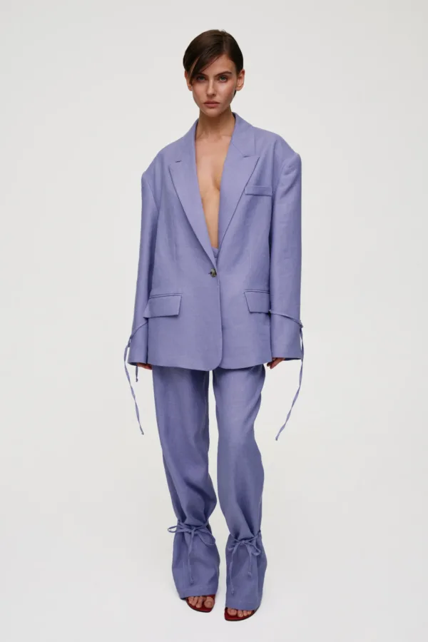 Woman wearing a lavender oversized suit with an open blazer revealing a bare chest, matching pants, and red sandals against a light background.