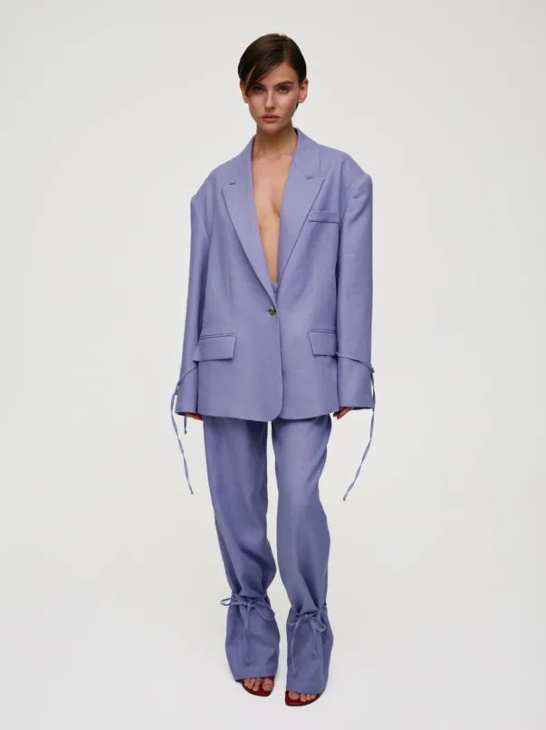 Woman wearing a lavender oversized suit with an open blazer revealing a bare chest, matching pants, and red sandals against a light background.