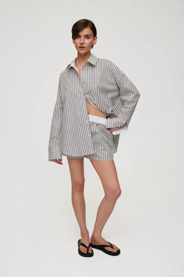 Woman standing in a relaxed pose, wearing a white and black vertical-striped oversized shirt paired with matching striped shorts and black flip-flops against a plain light backdrop.