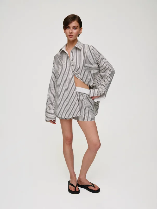 Woman standing in a relaxed pose, wearing a white and black vertical-striped oversized shirt paired with matching striped shorts and black flip-flops against a plain light backdrop.