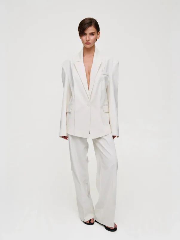 Model wearing an oversized white suit with an open blazer and white trousers, standing against a light background in a studio.
