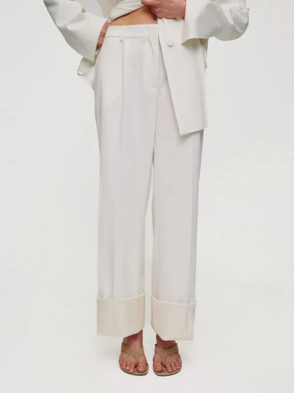 Lower half of a person in a white tailored suit with cuffed wide-leg pants and beige sandals.