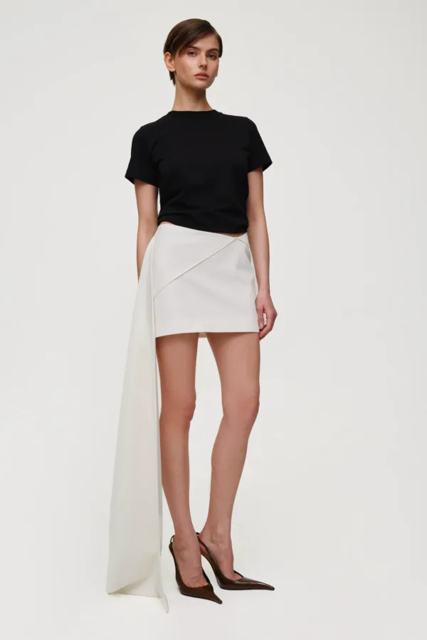 Model wearing a black short-sleeve T-shirt with a white asymmetric wrap skirt and brown heels, standing against a light background.