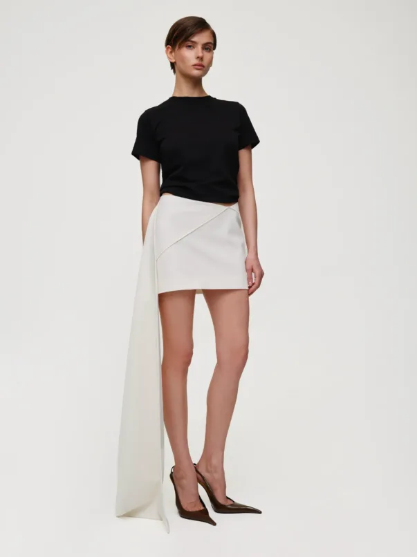 Model wearing a black short-sleeve T-shirt with a white asymmetric wrap skirt and brown heels, standing against a light background.