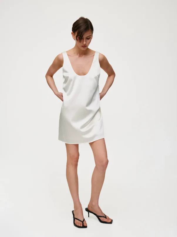 Model wearing a white satin sleeveless mini dress with a deep scoop neckline, standing with hands on hips against a white studio backdrop.