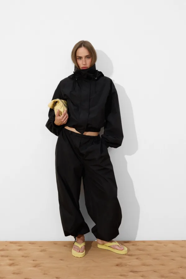 Person in a black windbreaker and pants stands against a white wall, holding a small yellow bag and wearing yellow sandals.