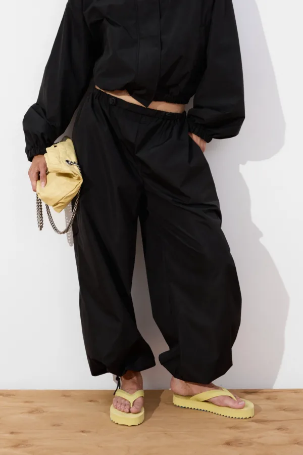 Person in black cropped top and loose black pants stands on a wooden floor, holding a small yellow quilted clutch with a chain strap.