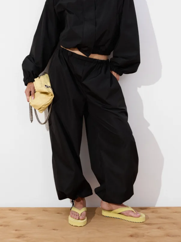 Person in black cropped top and loose black pants stands on a wooden floor, holding a small yellow quilted clutch with a chain strap.