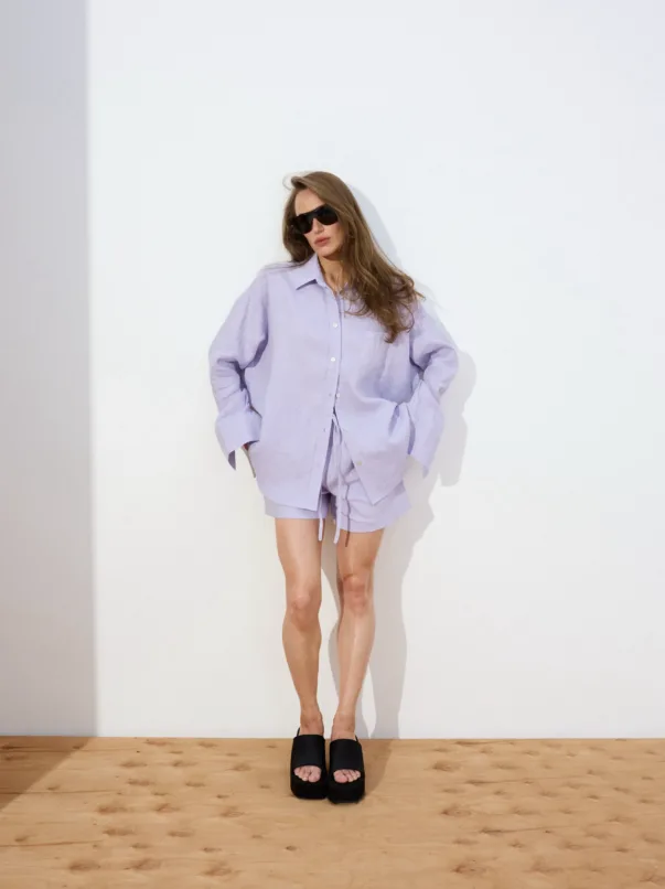 Woman in an oversized lavender shirt and matching shorts, wearing black sunglasses, standing with hands in pockets against a white wall on a wooden floor.