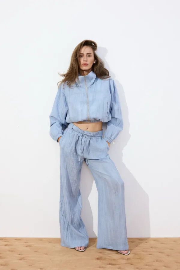 Model wearing a light blue matching jacket and wide-leg pants, standing against a white wall with a wooden floor, hands in pockets.