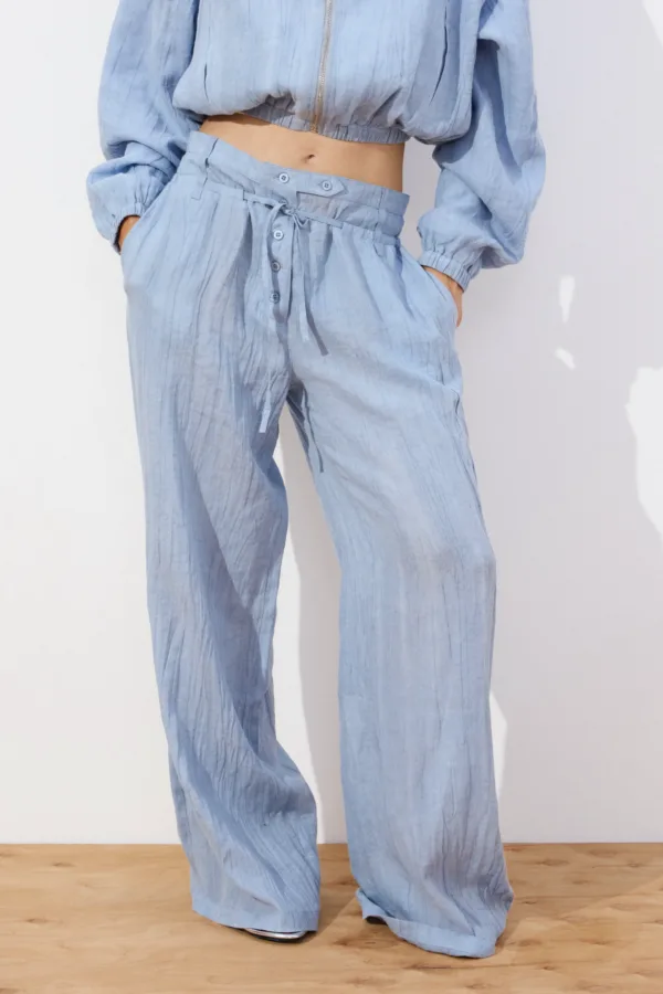 Person wearing light blue, loose-fitting pants with a drawstring waist and matching cropped top, standing against a white wall with hands in pockets.