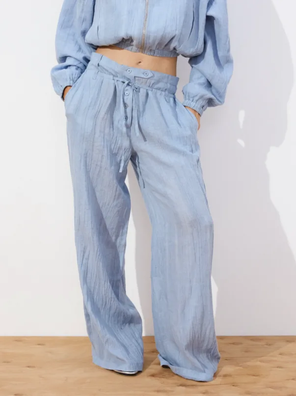 Person wearing light blue, loose-fitting pants with a drawstring waist and matching cropped top, standing against a white wall with hands in pockets.