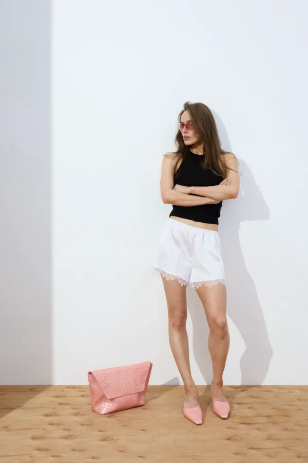 Woman standing against a white wall with arms crossed, wearing a black sleeveless top, white lace shorts, pink sunglasses, and pink flats; pink bag on the wooden floor nearby.