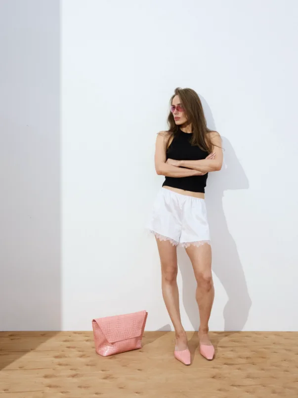 Woman standing against a white wall with arms crossed, wearing a black sleeveless top, white lace shorts, pink sunglasses, and pink flats; pink bag on the wooden floor nearby.