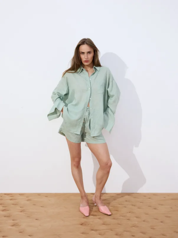 Woman in a sage-green oversized shirt and matching shorts stands against a white wall, hands in pockets, wearing pink heeled mules.