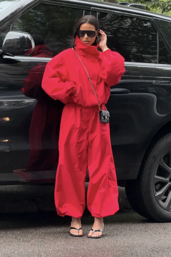 Person in a bright red jumpsuit standing in front of a black SUV, wearing oversized sunglasses and black sandals with a small black quilted purse on a chain strap across the body.