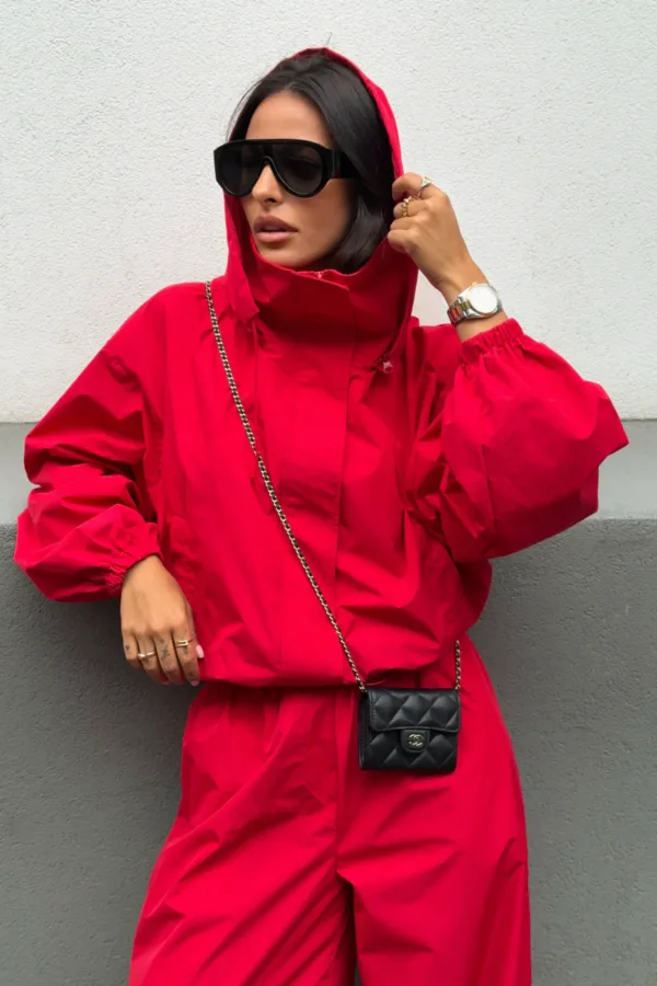 Woman in a red hooded jacket with oversized black sunglasses, posing against a gray wall with a small black quilted crossbody bag on a chain.