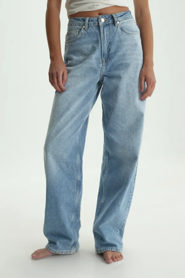Person wearing high-waisted blue jeans and a cropped white top, standing barefoot on a white background.